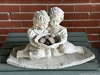 Front view of the cast cement garden sculpture showing two children sitting and holding a leaf-shaped basket filled with stones, placed on a wooden surface against a brick wall.
