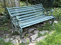Full angled side view showing green painted cast iron frame with lion head and scroll details, and wooden slats for seating and backrest. Positioned on irregular flagstone among grass and garden trellis backdrop.