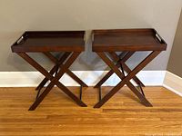 Pair of dark brown wooden serving trays on folding X-leg stands, positioned side by side against a wall.