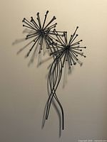 Metal wall art with two stylized flower structures made of black metal rods with spherical tips, showing side perspective on a cream wall.