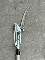 Close view of the metal cutting and pruner mechanism at the pole end with rope actuating the shear blades.