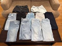 Top view of various folded girls pants and shorts on a dark table, includes denim jeans, sweatpants and shorts in light blue, black, gray and navy colors.