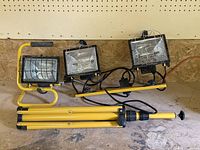 Set of two halogen work lights, one single light unit and one double light unit on yellow tripod stand with cords.