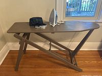 Wooden folding ironing board with two clothing irons placed on top, viewed from side near window and hardwood floor.