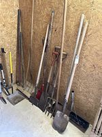 Photo showing various long-handled garden and outdoor tools leaning against a wood wall, including shovels, pitchfork, sledgehammer, garden forks, and a metal rake.