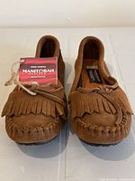 Front view of pair of brown suede Manitobah mukluks moccasins showing fringed trim and tags attached
