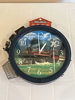 Front view of round quartz clock with steam train image and model train circling perimeter. 'Train Station' building at top center.