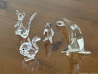 Four clear animal figurines on a wooden surface: Swarovski crystal squirrel, glass seal, glass penguin, and glass kangaroo.