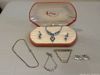 Full lot including blue rhinestone necklace set in original Calzena Jewelry box, gold-tone snake chain necklace, silver-tone bird pendant necklace, gold-tone tie clip with chain, and silver-tone cufflinks.