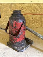 Side view of vintage hydraulic bottle jack showing worn red and black paint and metal handle.