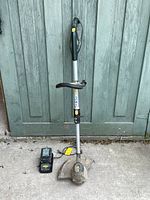 Full view of Yardworks cordless string trimmer with attached curved shaft, telescopic handle, battery, and charger placed on concrete ground against green wooden door background.