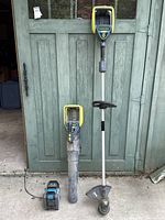 Full view of Yardworks cordless string trimmer and leaf blower with battery and charger on cement floor in front of green door.