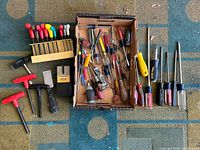 Overview of approximately 20 assorted screwdrivers and other hand tools laid out on a carpeted floor, including various handle colors and sizes.