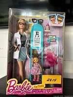 Front view of Barbie Eye Doctor playset in original packaging showing Barbie doll, child doll, eye exam chair, eye chart, and 4 pairs of glasses.