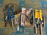 Tools laid out on carpet including loppers, saws, hammers, hand clamp, miter box, square, drill bits, and garden stakes