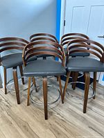 Five contemporary counter stools with brown wooden frames, dark grey cushioned swivel seats, and chrome metal footrests, arranged in a cluster indoors.