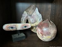 Three abstract decorative pieces on a shelf including a broken spherical pot, a wooden eyes sculpture, and a stone figure.