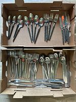 Two open cardboard boxes showing large sets of everyday flatware and serving pieces made of stainless steel. One set has twisted handles, the other set has plain handles.
