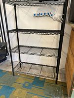 Photo showing the entire five-tier shelving unit made of black steel wire, positioned against a wall with some background objects.