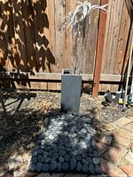 Full view of metal palm tree sculpture in planter pot placed on ground outside by a wooden fence, door mat with rounded stones in front