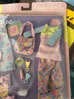 Front view of Midge Happy Family playset in original packaging showing doll clothes, including a pink and yellow dress over blue long-sleeve top, patterned pants, handbag, baby bottle, pacifiers, bib, and bootie.