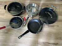 Overview of six cookware pieces on floor including pots and pans with lids, made by All-Clad and Masterclass.