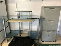 Photo showing the glass and metal computer desk with wooden keyboard tray and adjacent gray filing cabinet with three drawers.