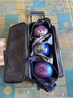 Roller bag unzipped with four different colored and branded bowling balls inside, showing arrangement and bag compartments.