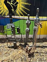 Photo showing four metal sculptures outdoors in front of a painted fence with mural, including abstract humanoid figures and geometric shapes.