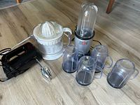 Photo shows Magic Bullet blender base, four clear plastic blending cups with handles placed around it, and a white manual juicer on wooden floor.