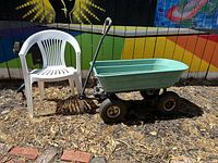 Outdoor garden cart and white plastic chair shown side by side on ground with dry grass and leaves, colorful painted fence in background