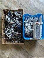 Two cardboard boxes containing a tangled assortment of black, white, and blue power cords, a white power strip with four outlets, plug adapters, and timer switches.