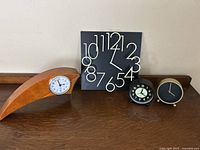 Four unique artistic clocks displayed on a wooden surface against a white wall, showing details and relative size of each clock.