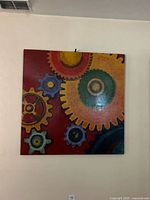Square painted canvas of abstract colorful gears on red and yellow background hung on a wall.