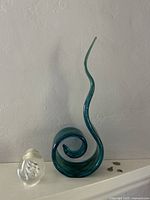 Photo showing blue glass curved sculpture and clear glass paperweight together on a white surface against a textured wall.