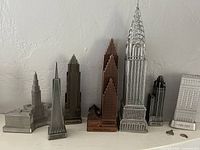Front view of seven metal models of famous American skyscrapers arranged in a row on a shelf against a white wall. Models include the Chrysler Building and others in silver and bronze finish.