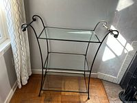 Full view of the three-shelf glass and iron cart near window showing top and middle glass shelves and black iron frame with curved handles.