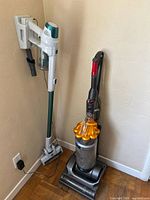 Dyson upright vacuum alongside Kenmore CSV Go stick vacuum mounted on wall mount in corner.
