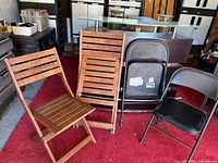 Four folding chairs: two wooden slat folding chairs in natural brown finish, two black metal folding chairs. Chairs shown standing on red carpet in garage setting.