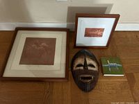 Two framed brown-toned artworks, a tribal wooden mask, and a decorative small green book arranged on a wood floor