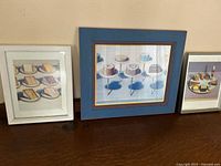 Three framed prints side by side: left white frame with cake slices, center blue frame with cakes on stands, right silver frame with pastries