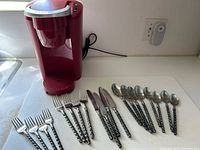 Red Keurig coffee maker with stainless steel flatware displayed on countertop. Flatware includes forks, knives, and spoons with twisted style handles.