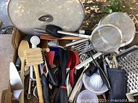 Assorted kitchen tools and utensils in a box, including wooden spoons, graters, spatulas, peelers, citrus juicers, strainers, knives, and more.