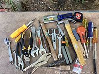 Overview of assorted small hand tools including wrenches, pliers, hammers, scissors, and screwdrivers spread on a concrete surface.