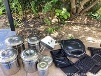 Overview photo showing the stainless steel canisters arranged in descending sizes, the hand mixer, waffle iron, and sandwich press plates outdoors on a table under shade.