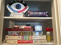Shelf view showing Eye Know trivia game shaped like an eye, Outburst II, Payday, The Game of Life, Monopoly Anniversary Edition, Scrabble, and more stacked games