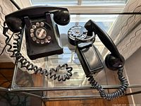 Photo showing the pair of black rotary dial telephones, one fully assembled, the other with handset detached. Coiled cords and rotary dials visible on both.