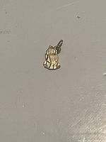 Photo of a small 10k gold charm shaped like praying hands, shown on a flat surface with visible details of the fingers and loop for attachment.