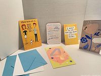 Five assorted greeting cards standing or lying on a white surface with blue envelopes, showing varied designs and messages.