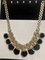 Front view of large gold-tone chain necklace with nine black geometric stone-like pendants showing glossy finish and segmented link design.